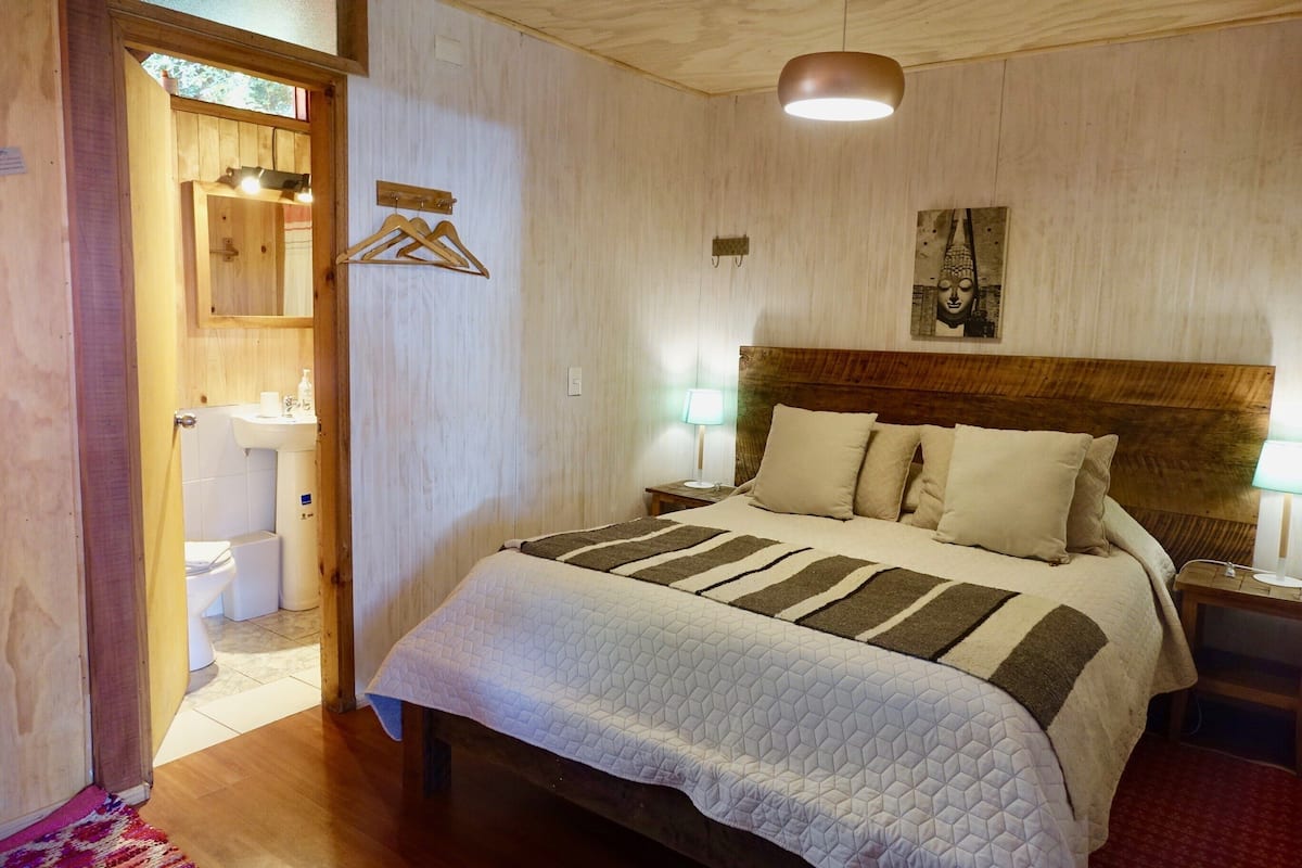 A bedroom with a wooden headboard, a bed with a striped blanket, a nightstand with a lamp, and a wooden door leading to a bathroom.