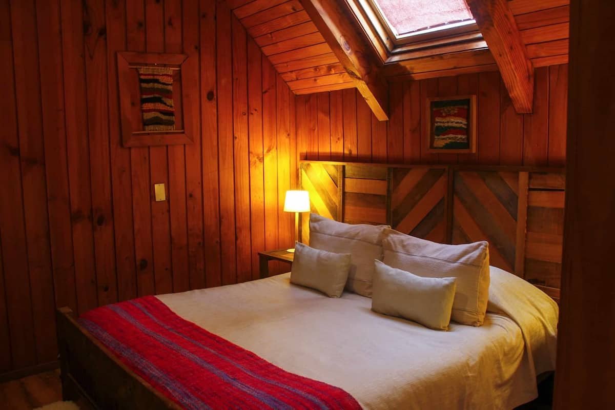 A wooden cabin bedroom with a bed, pillows, a nightstand, a lamp, and a framed picture on the wall.