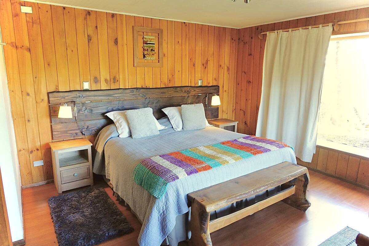 A wooden-paneled bedroom with a bed, two bedside tables, a bench, and a window with curtains.