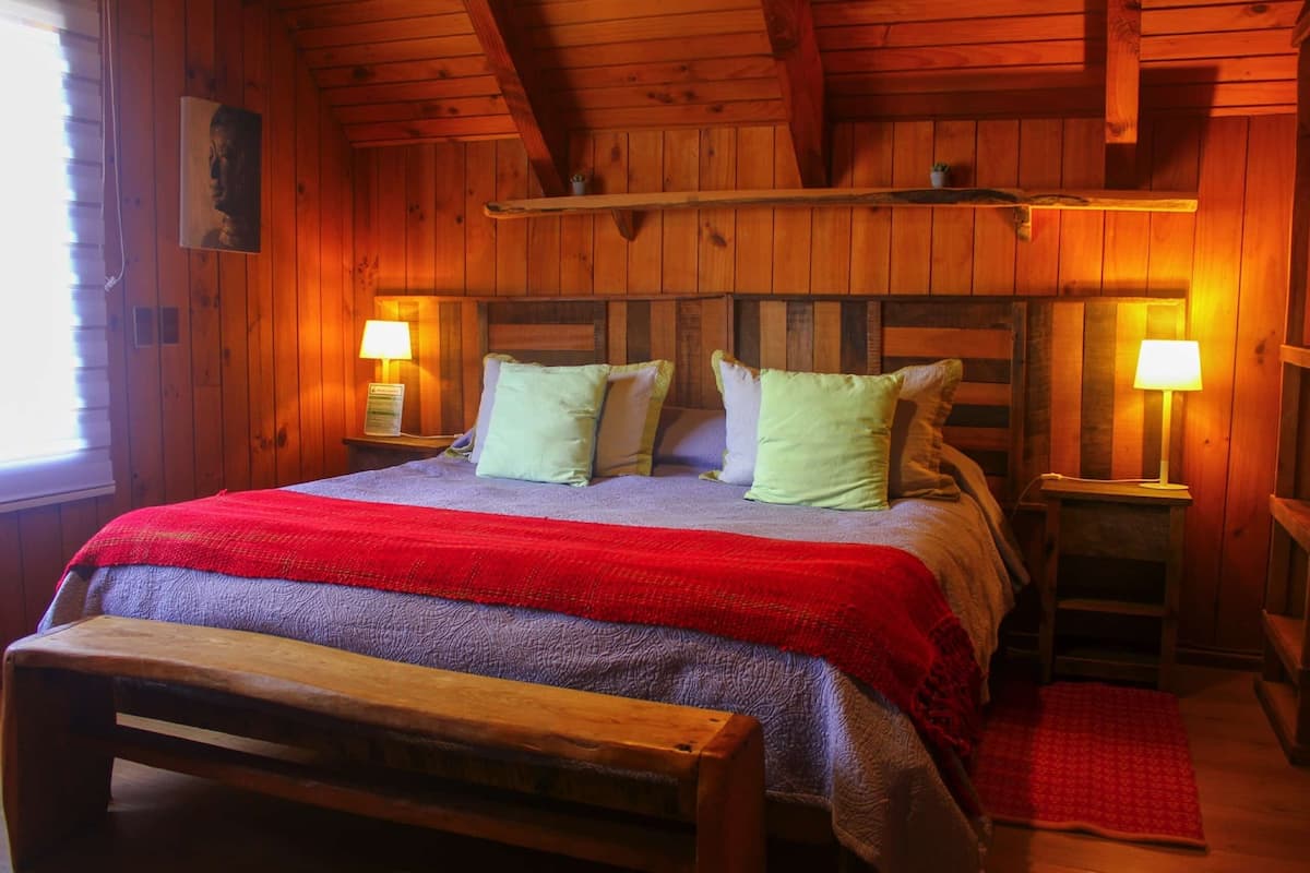 A wooden cabin-style bedroom with a bed, two bedside lamps, a bench, and a red rug.