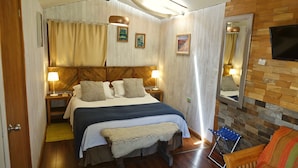 Un dormitorio con cabecera de madera, dos mesitas de noche con lámparas, un banco, una silla y un televisor montado en la pared.