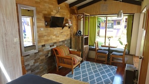 Una habitación acogedora con pared de madera, un televisor sobre una estantería, una silla de madera con un cojín colorido, un tapete estampado y una mesita con una planta.