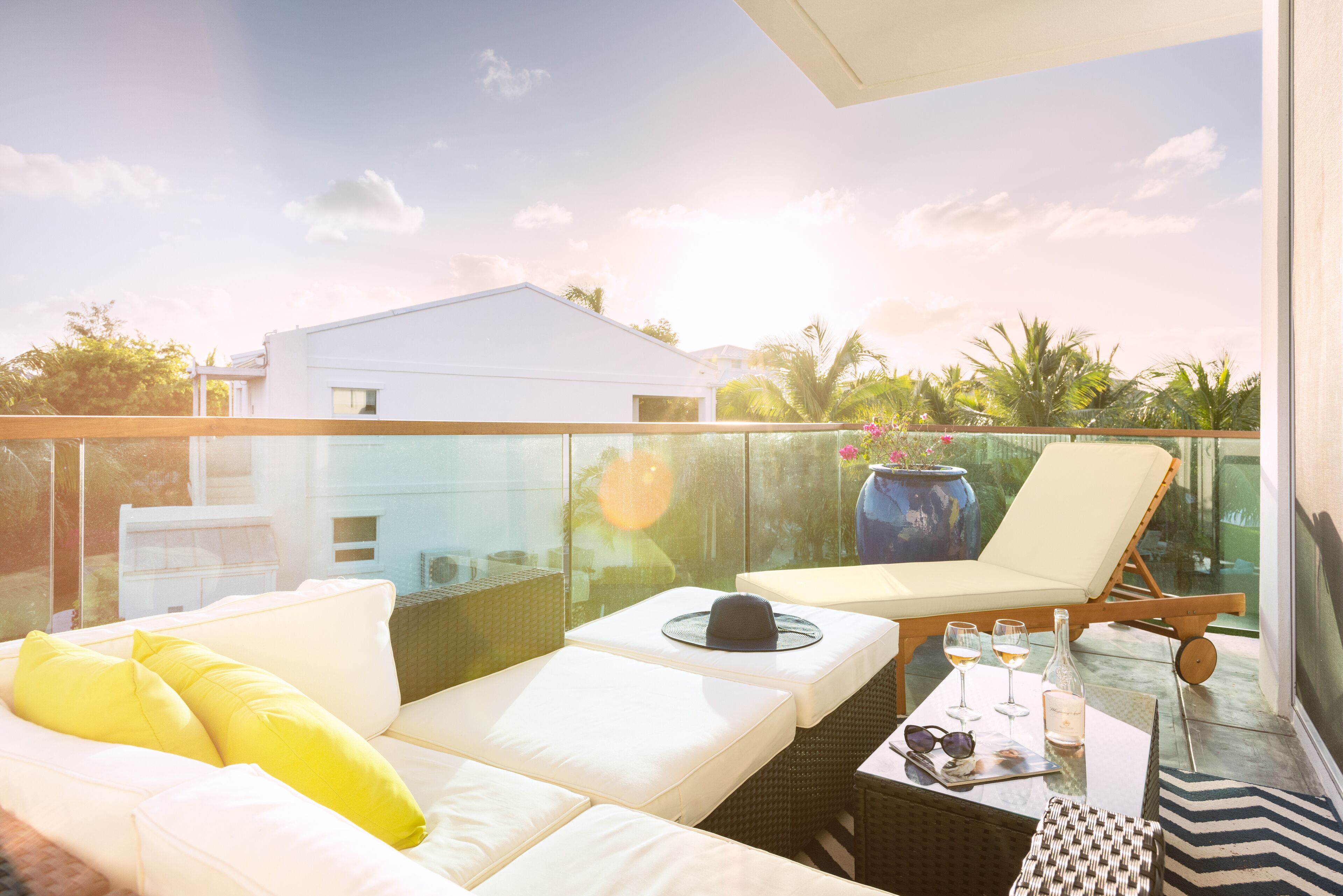 A balcony with a white sofa, yellow cushions, a wicker table with a hat, wine glasses, and a vase.