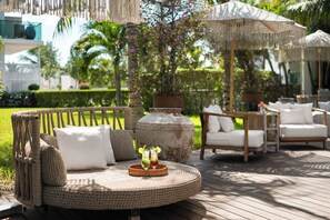A round wicker sofa with cushions and a woven tray with drinks, surrounded by wicker furniture and umbrellas, set on a wooden deck with a garden and palm trees in the background.