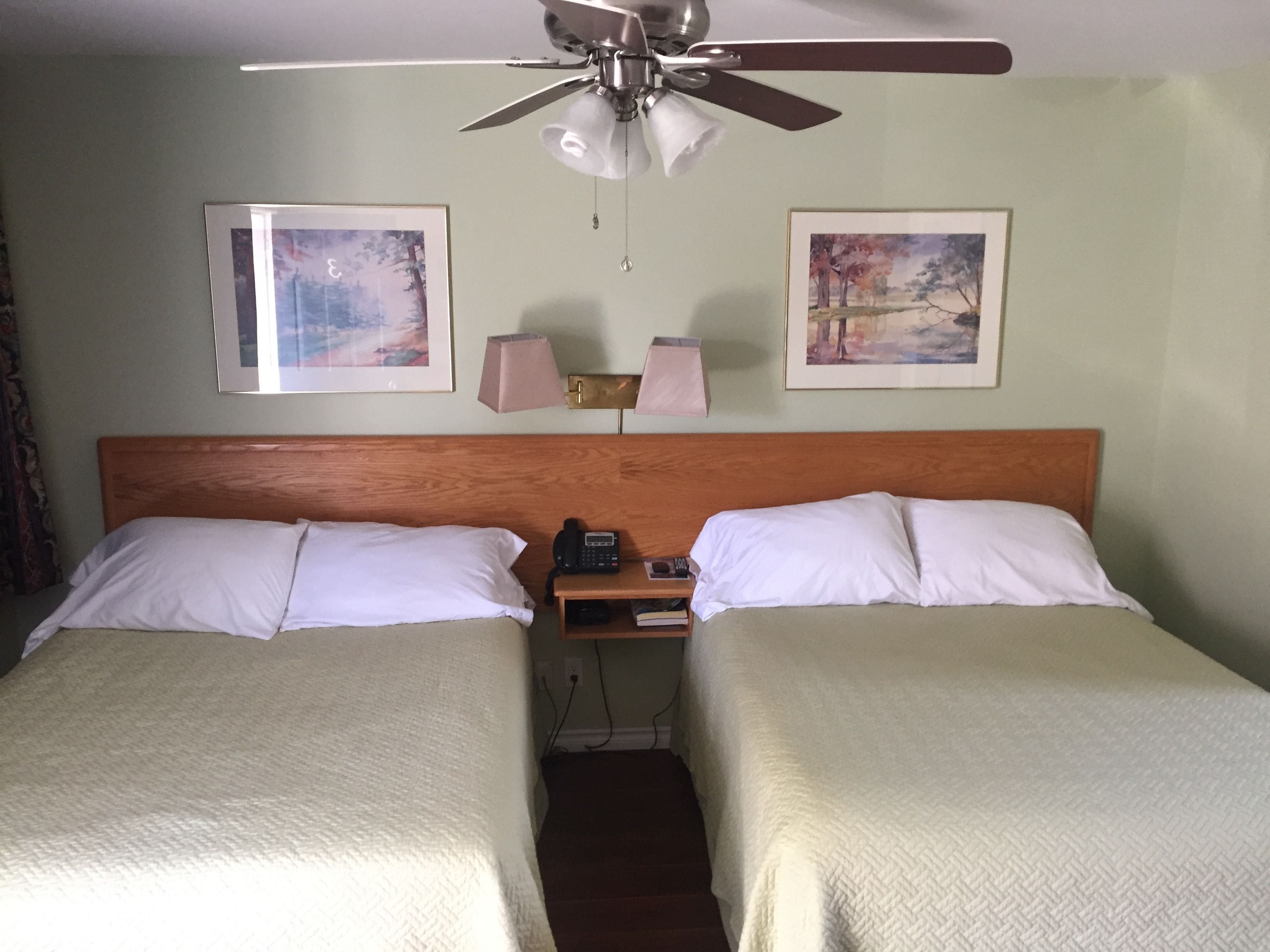 A hotel room with two single beds, a ceiling fan, two bedside lamps, and two framed pictures on the wall.
