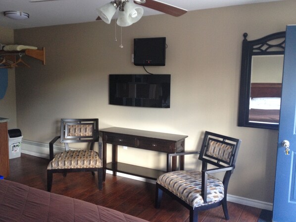 A room with a ceiling fan, a TV mounted on the wall, a wooden desk, two chairs, and a door.