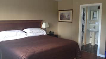 A hotel room with a large bed, a nightstand, a lamp, a painting on the wall, and a bathroom visible through an open door.