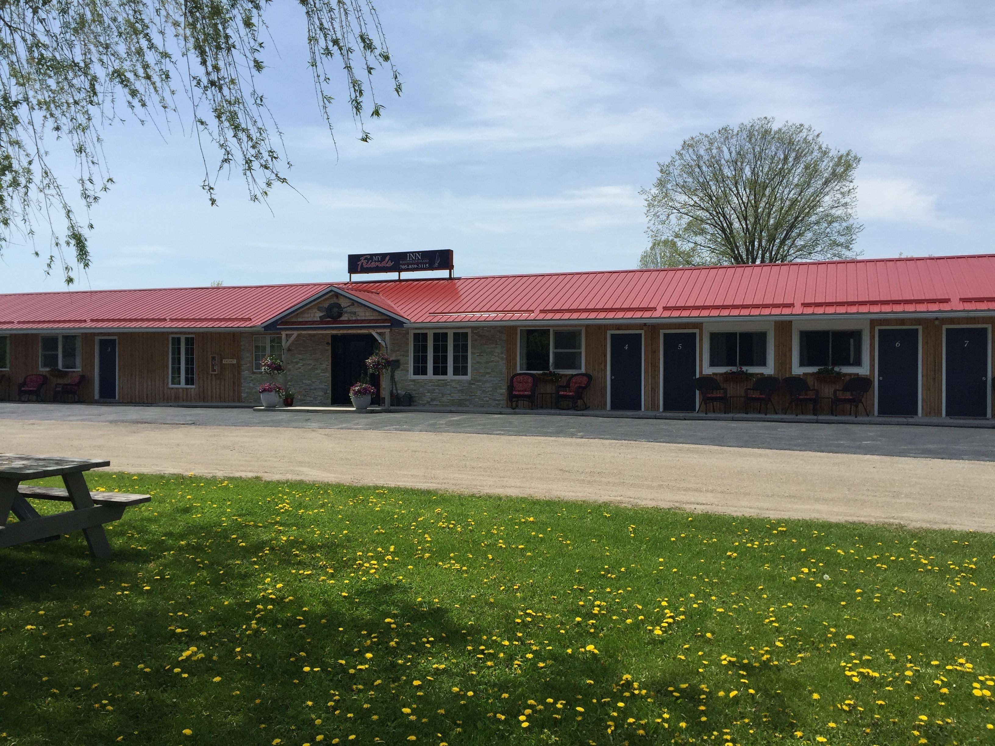 Un motel con techo rojo y varias habitaciones, un área de estacionamiento techada y una mesa de picnic al frente.