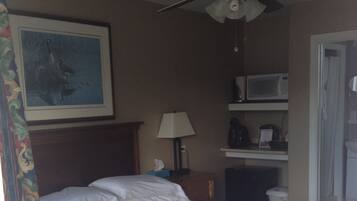 A bedroom with a bed, a ceiling fan, a painting on the wall, a lamp, and a microwave on a shelf.