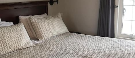 A neatly made bed with a quilted cover, a wall-mounted lamp, and a window with curtains.
