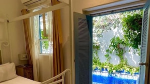 Une chambre dotée d’un lit à baldaquin, d’un climatiseur, d’une fenêtre avec des rideaux et d’une porte ouverte donnant sur un espace piscine.