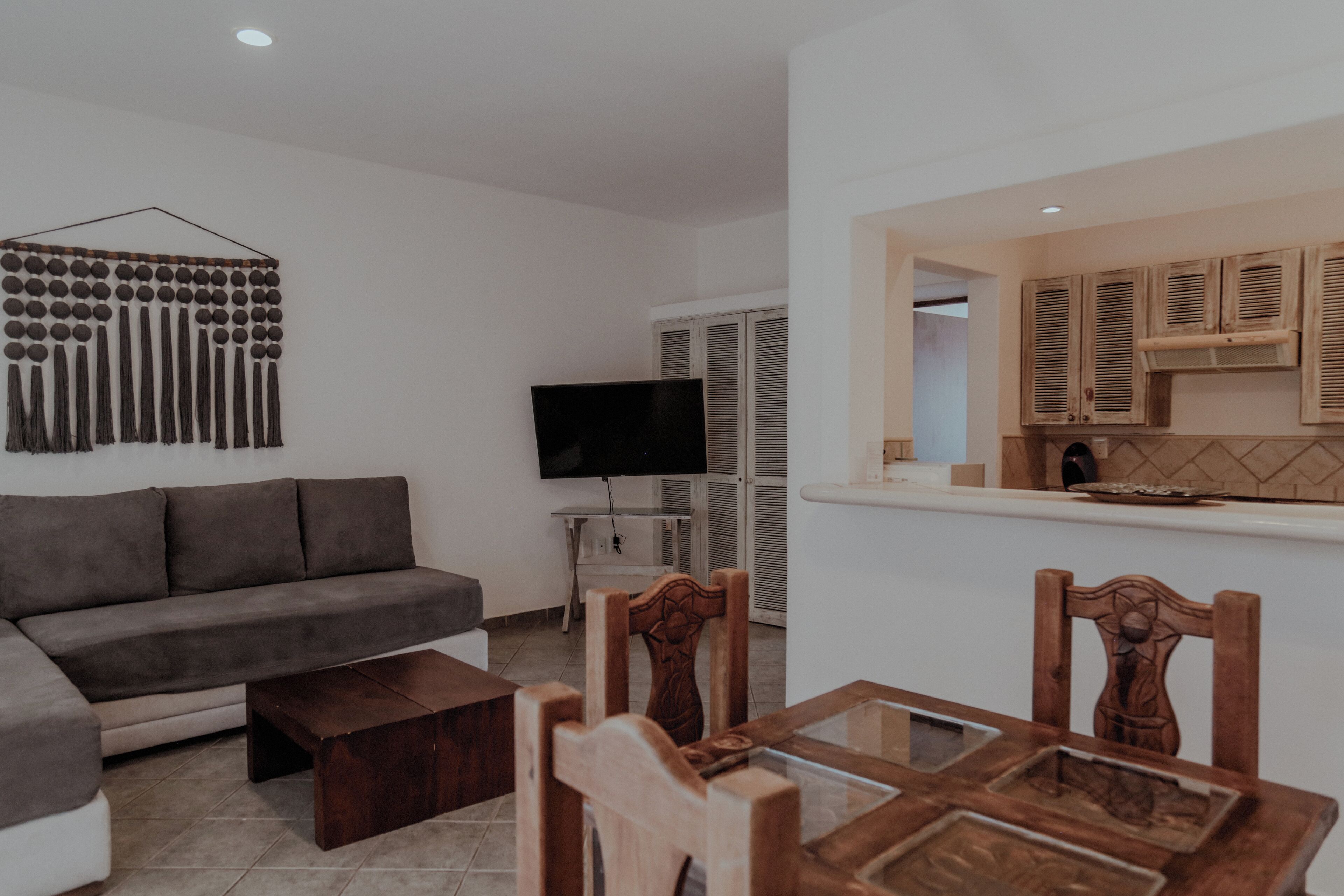 A modern living room with a grey sofa, wooden dining table, and chairs. A flat-screen TV is mounted on the wall.