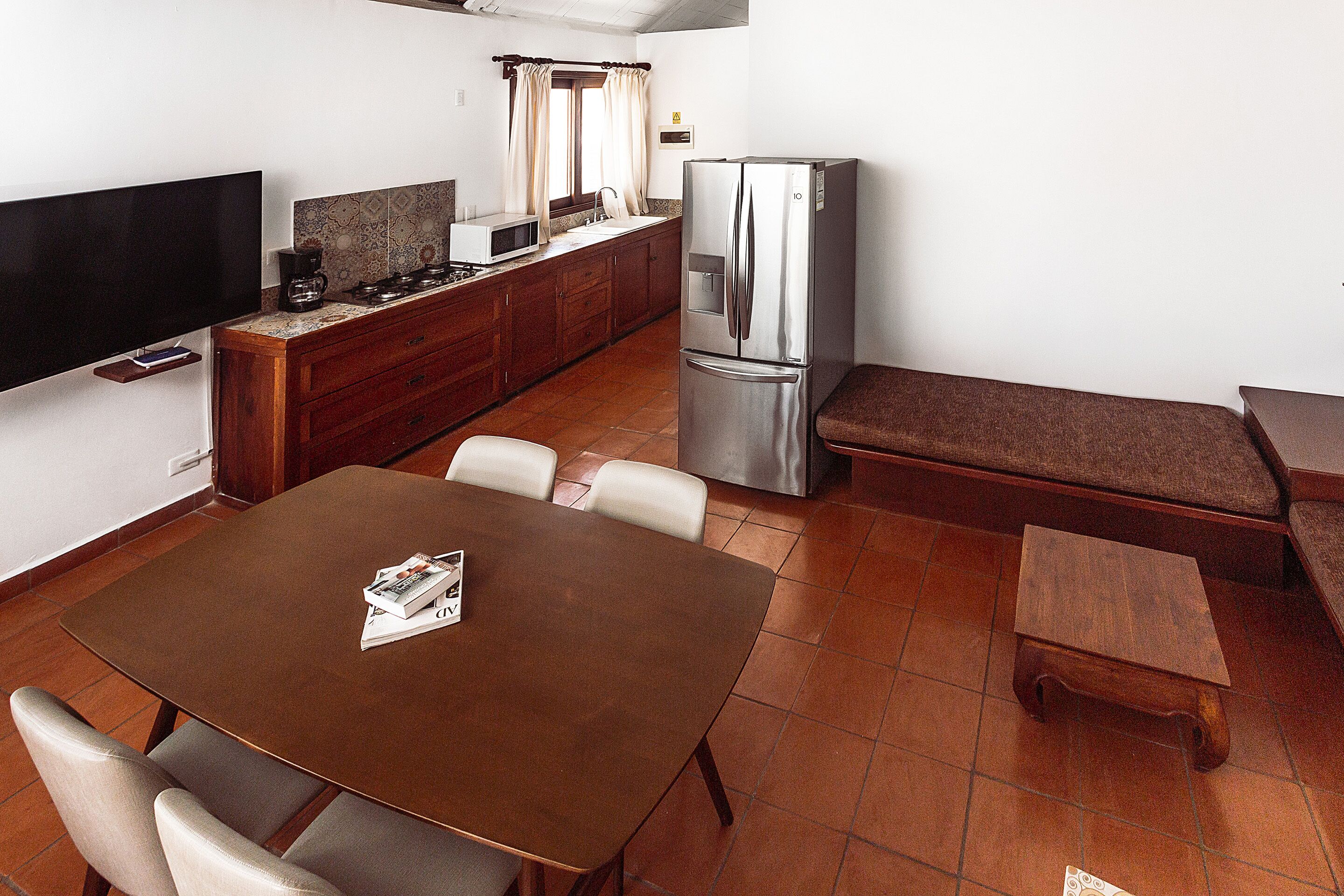 A compact kitchen with a refrigerator, microwave, and a flat-screen TV. There is a dining table with chairs and a small bench.