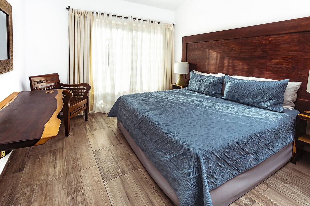 Ein Hotelzimmer mit einem großen Bett, einem Nachttisch aus Holz, einem Sessel, einem Holztisch und einem Fenster mit Vorhängen.