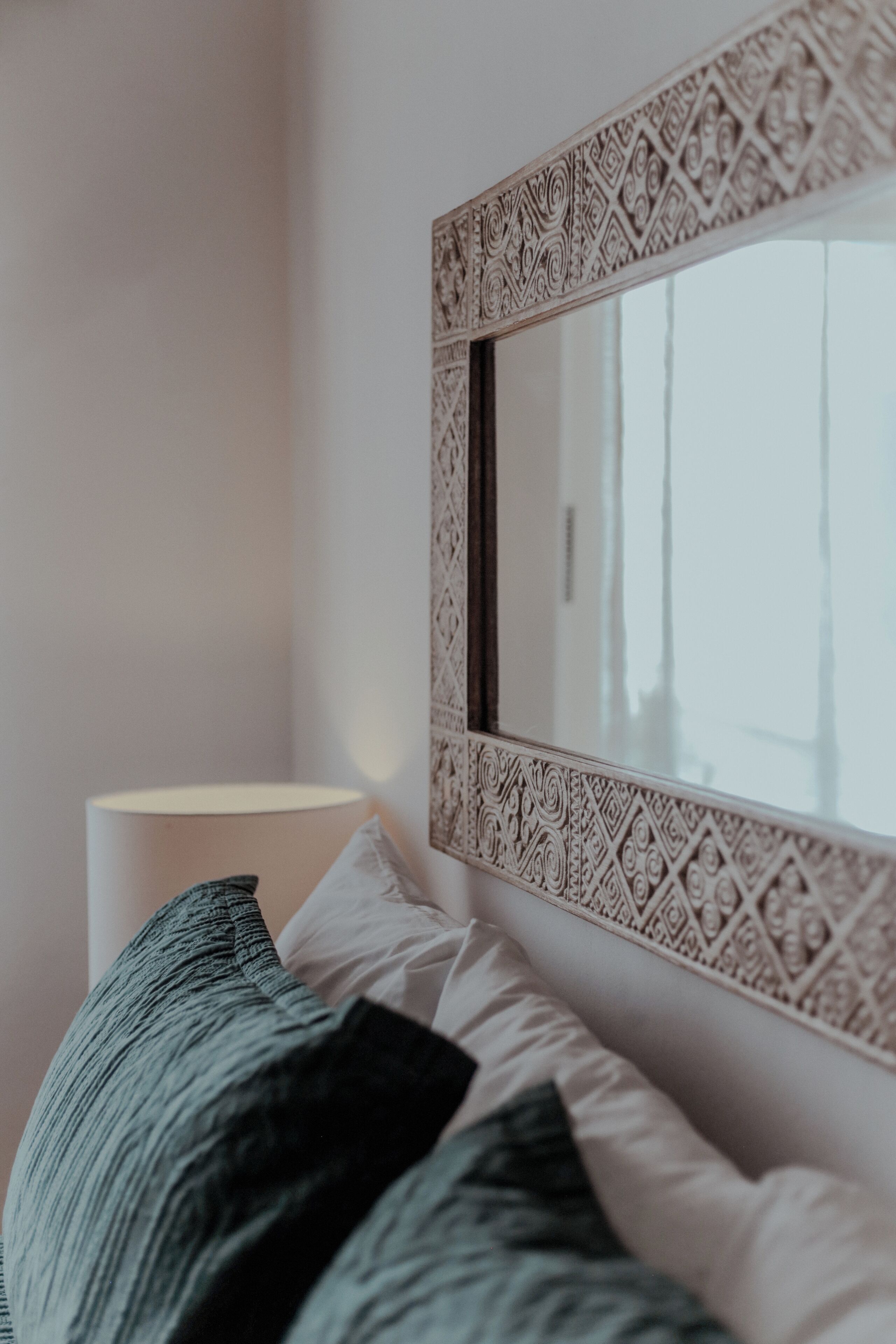 Un dormitorio con cama, almohadas, una lámpara y un espejo decorativo con un marco detallado.