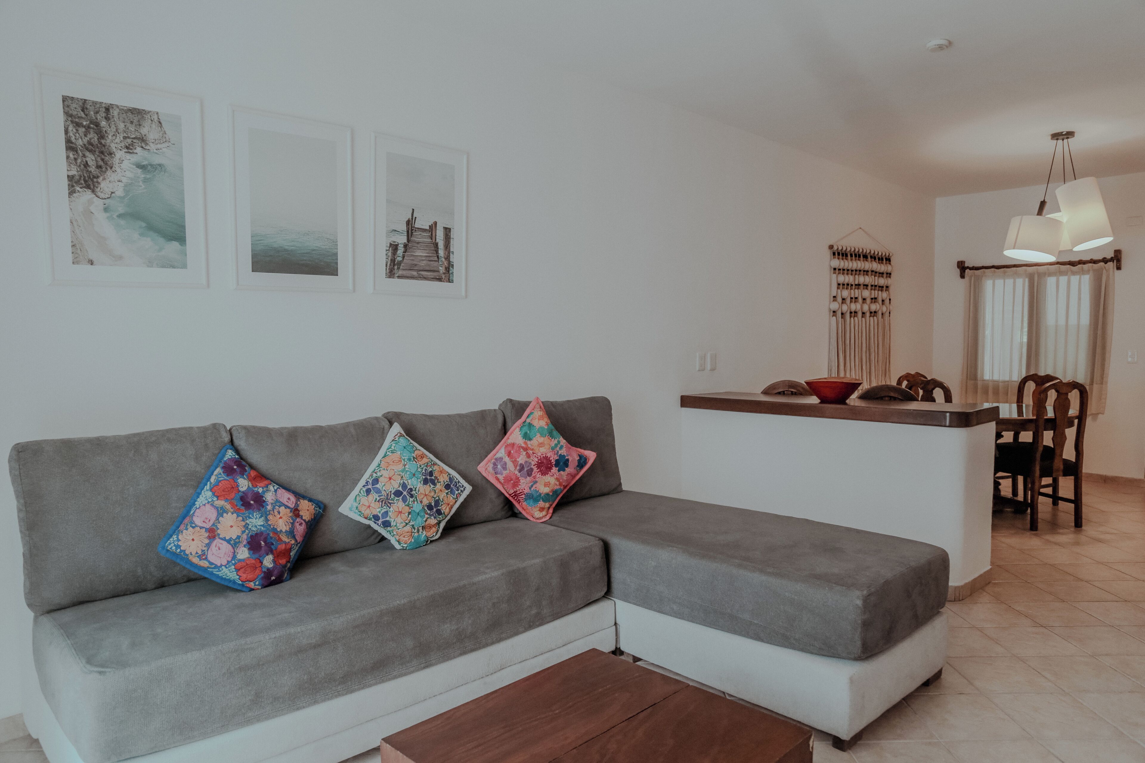A modern living room with a grey sectional sofa, colorful floral cushions, a small kitchen area, and a dining table with chairs.