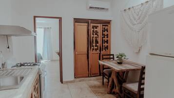 Una cocina con estufa a gas, un lavamanos y una mesa de madera con sillas. Hay un armario de madera con puerta decorativa y un colgador de macramé en la pared.