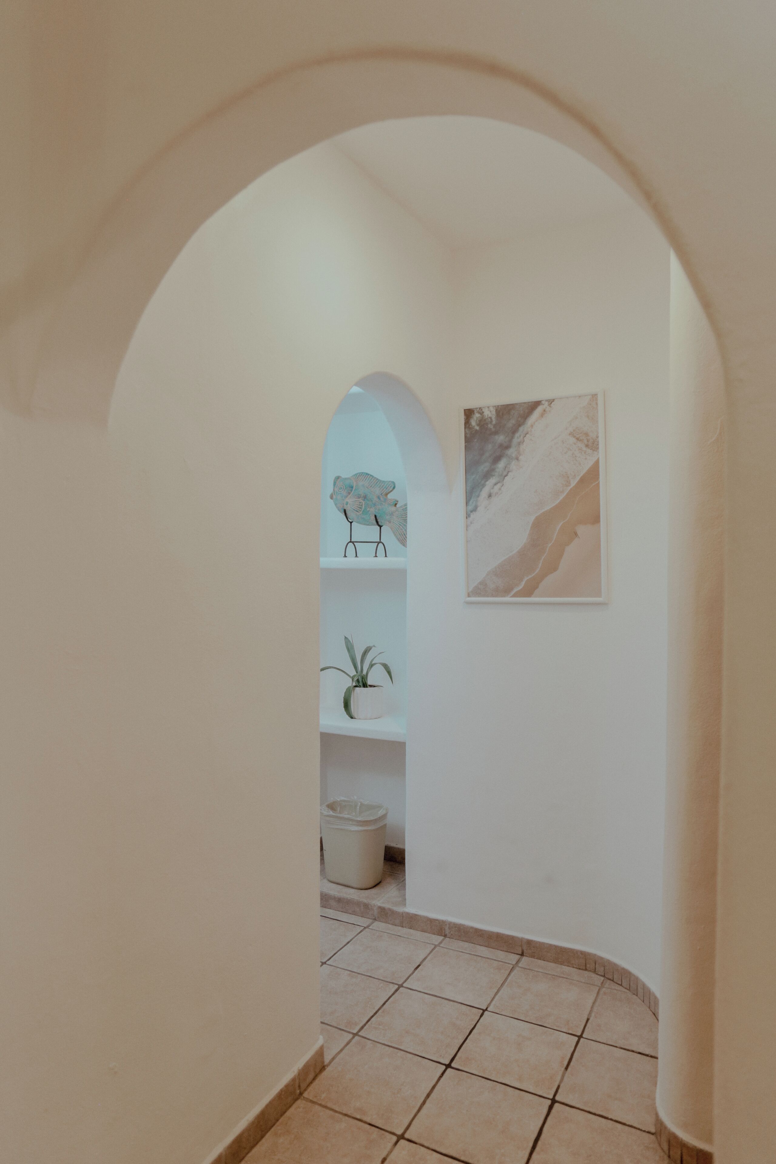A hallway with a tiled floor, a white wall with a framed picture, and a shelf with decorative items.