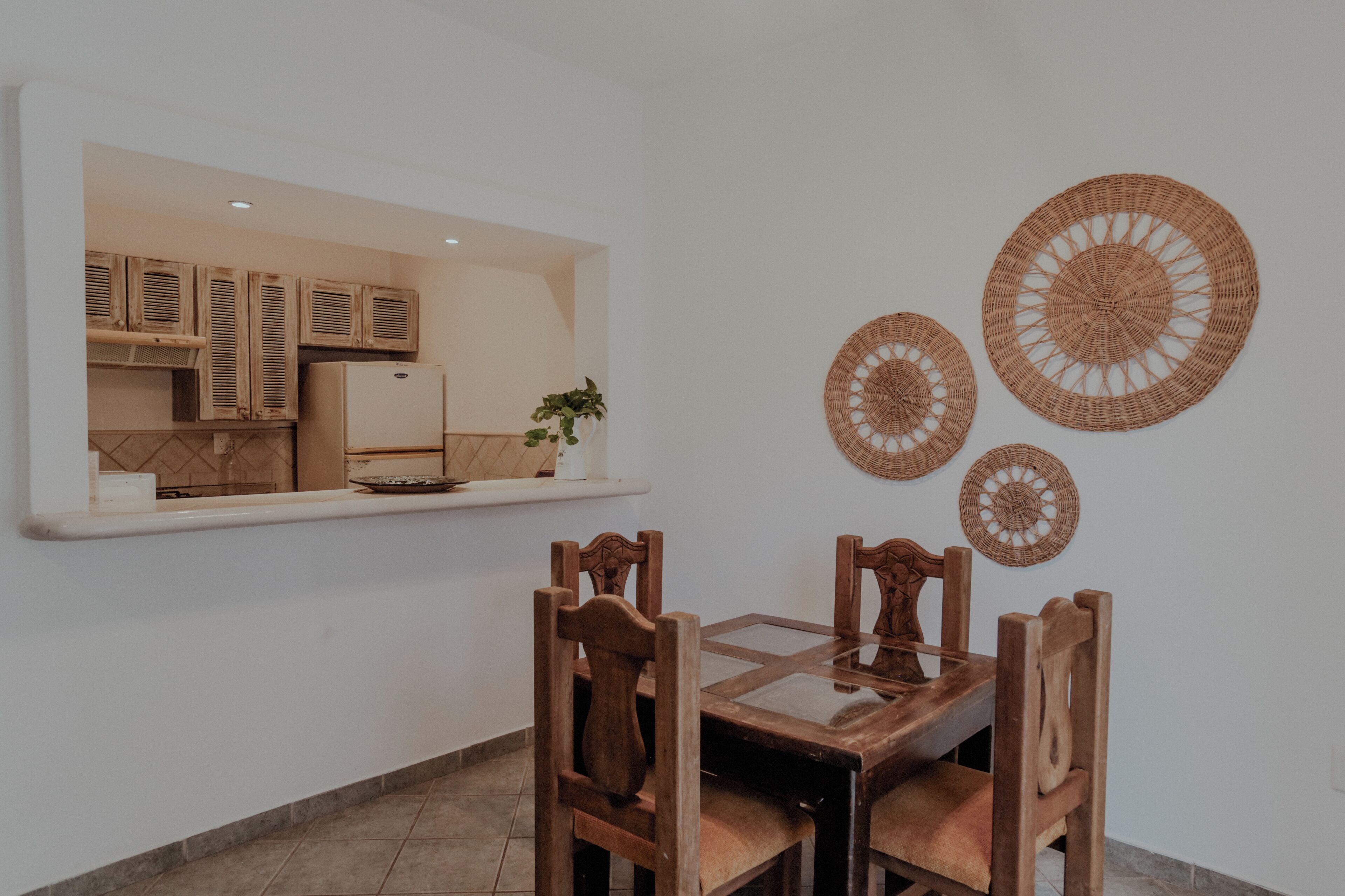 A dining area with a wooden table and chairs, a kitchen visible through an open door, and three woven wall decorations.