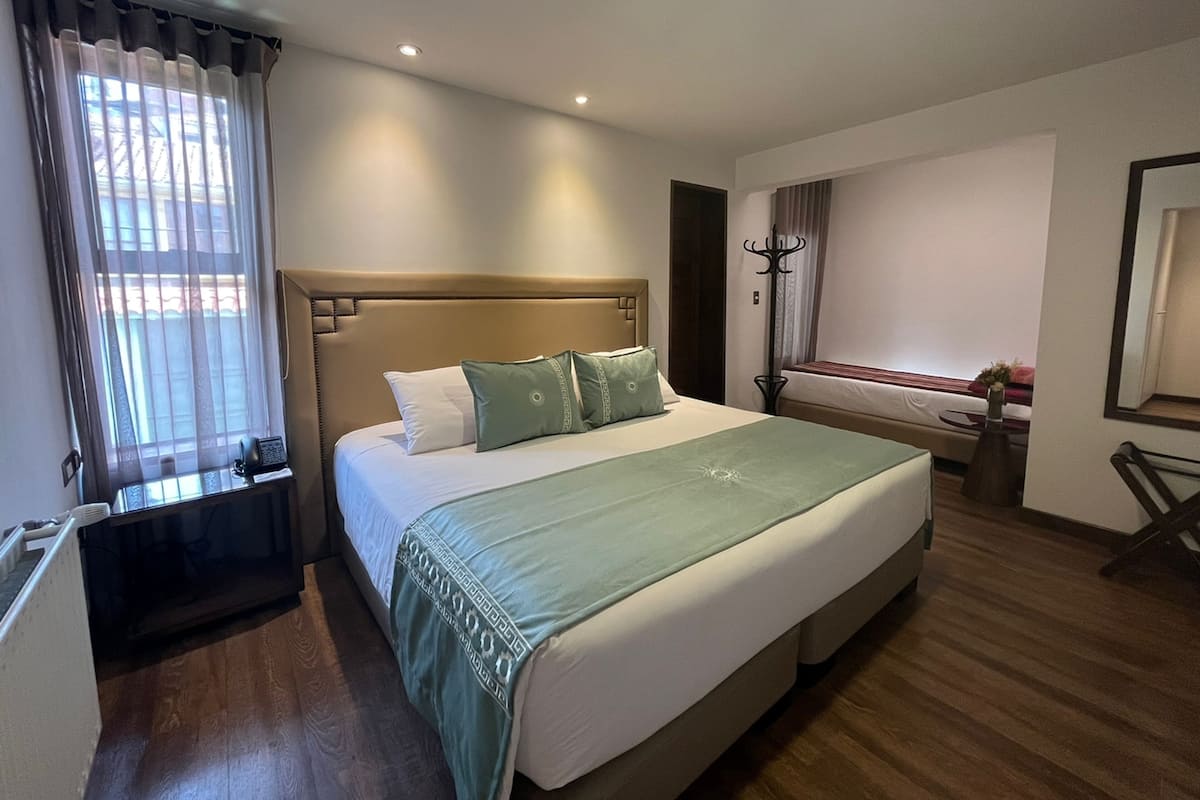 A hotel room with a large bed, a bedside table, a window with curtains, a coat rack, and a mirror on the wall.