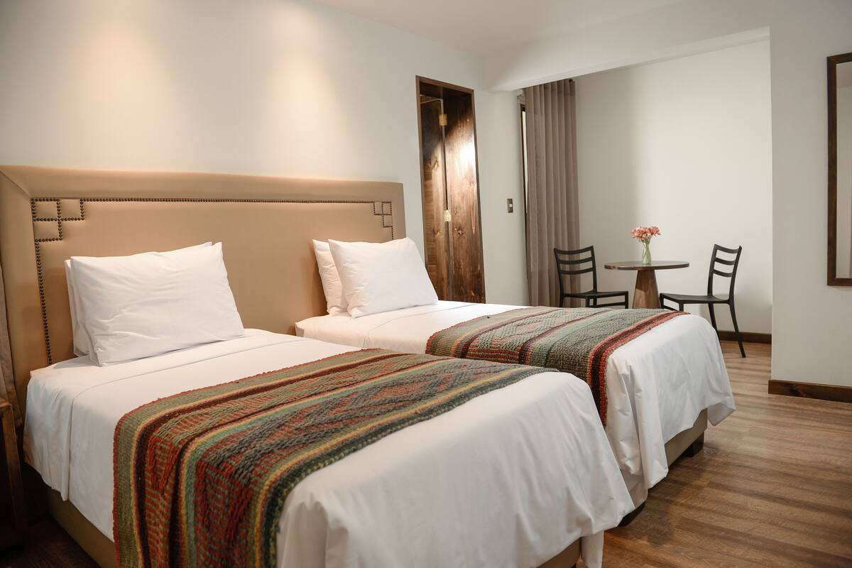 A hotel room with two beds, a wooden headboard, a small table with chairs, and a window with curtains.