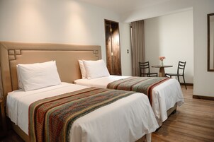 Habitación de hotel con dos camas, un cabecero de madera, una mesita con sillas y un ventanal con cortinas.