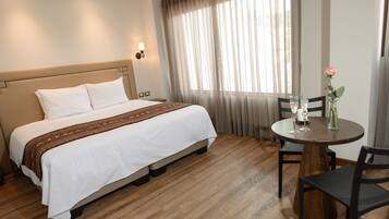 A hotel room with a bed, a round wooden table with two chairs, a vase with a single flower, and a window with sheer curtains.