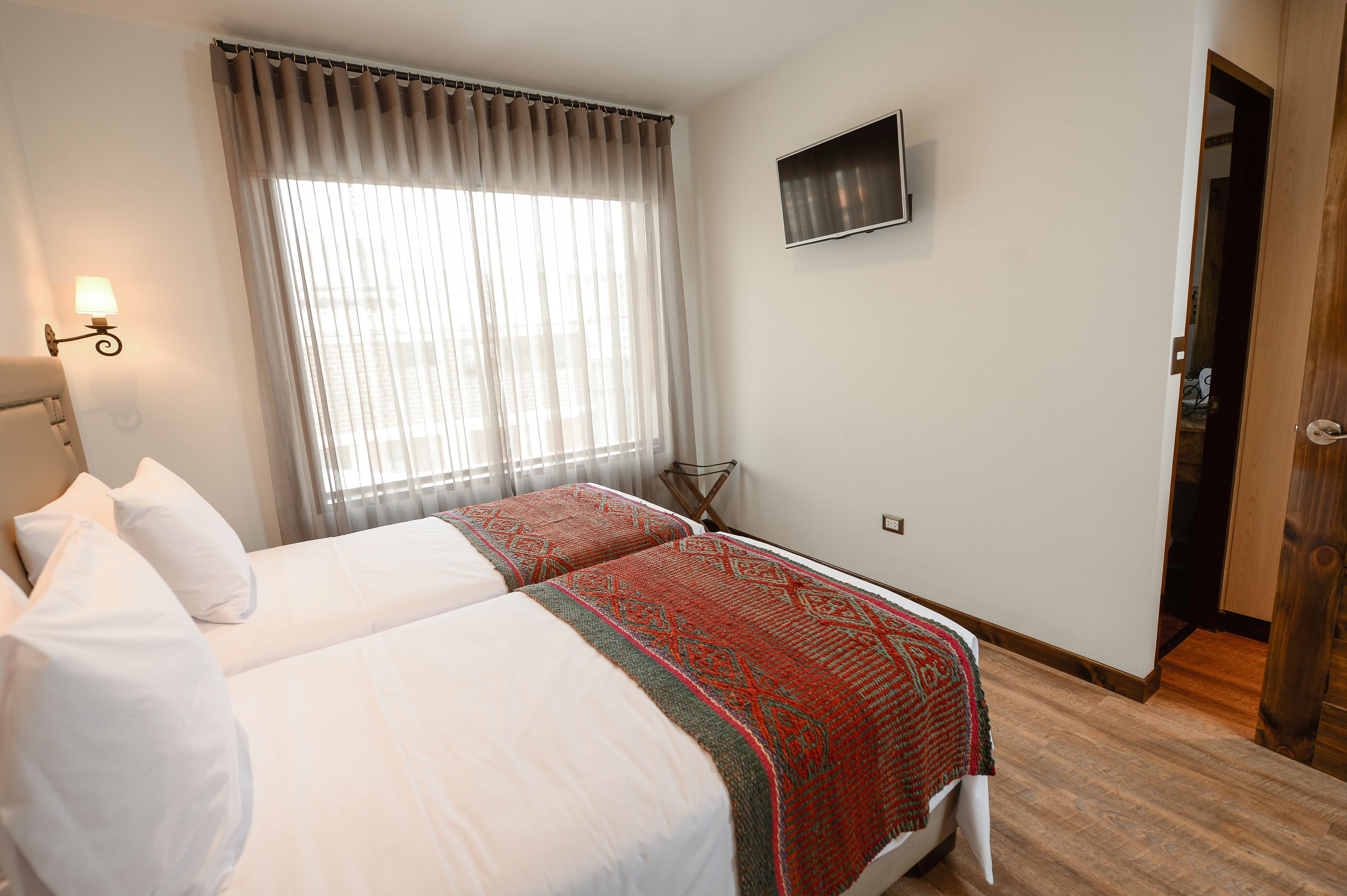 Habitación de hotel con dos camas, un televisor montado en la pared y un ventanal con cortinas.