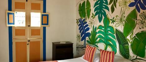 A bedroom with a bed, a nightstand, a wall with a tropical leaf mural, and a door with a blue frame.