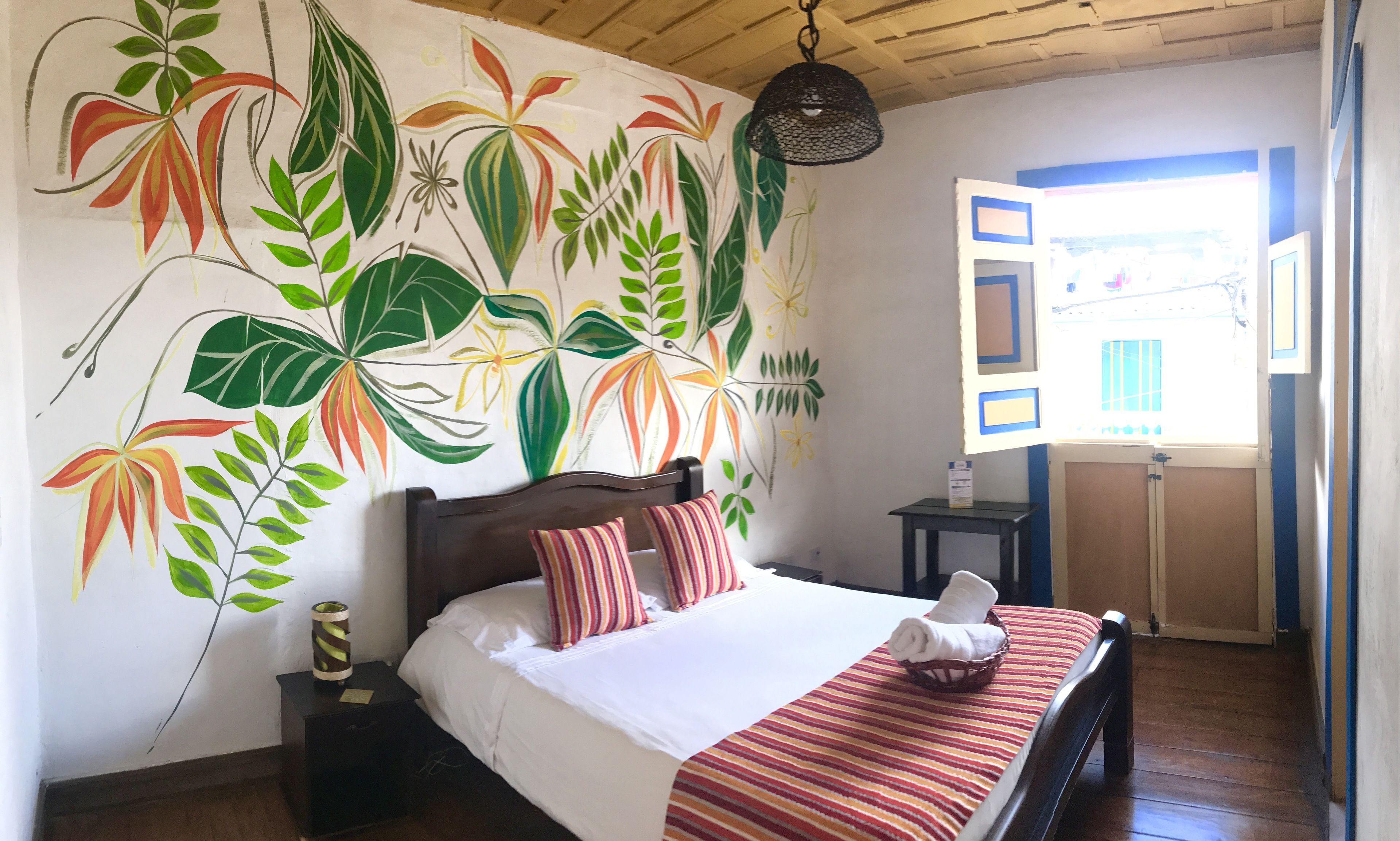 A bedroom with a bed, striped pillows, a nightstand, a lamp, and a wall mural of tropical plants.