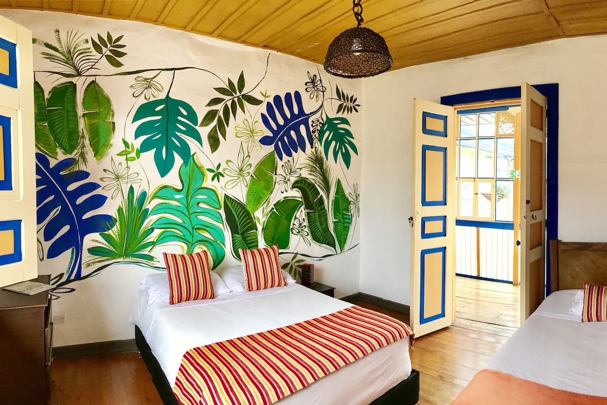 A room with two beds, a mural of tropical leaves on the wall, and a wooden ceiling.