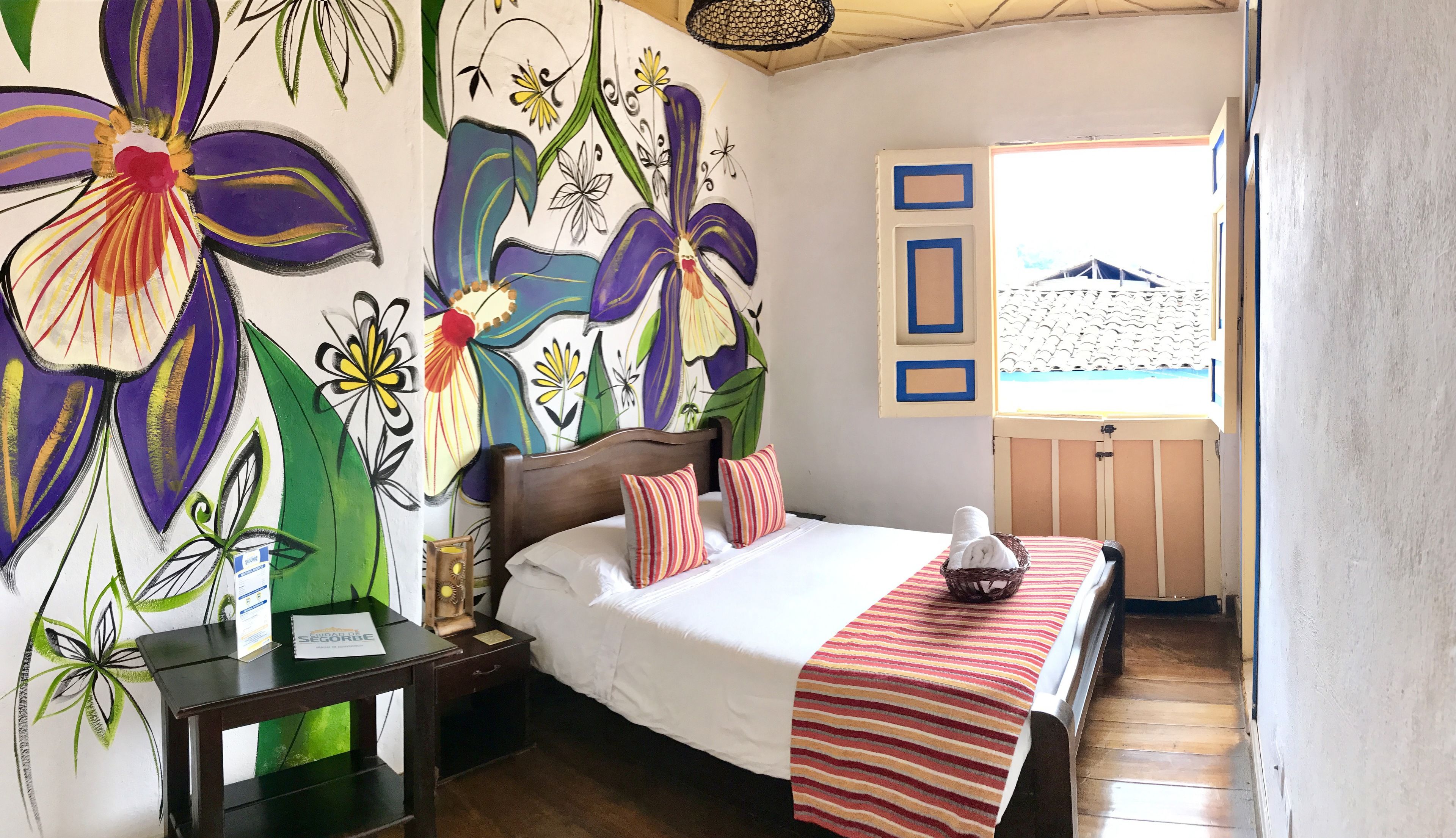 A bedroom with a large floral mural, a wooden bed, a nightstand, a desk, and a window with a view of a building.