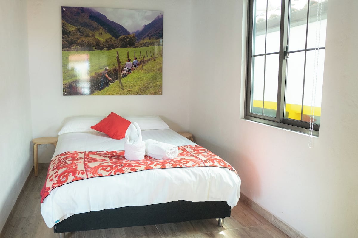 A hotel room with a bed, a red pillow, and a painting of a landscape on the wall.
