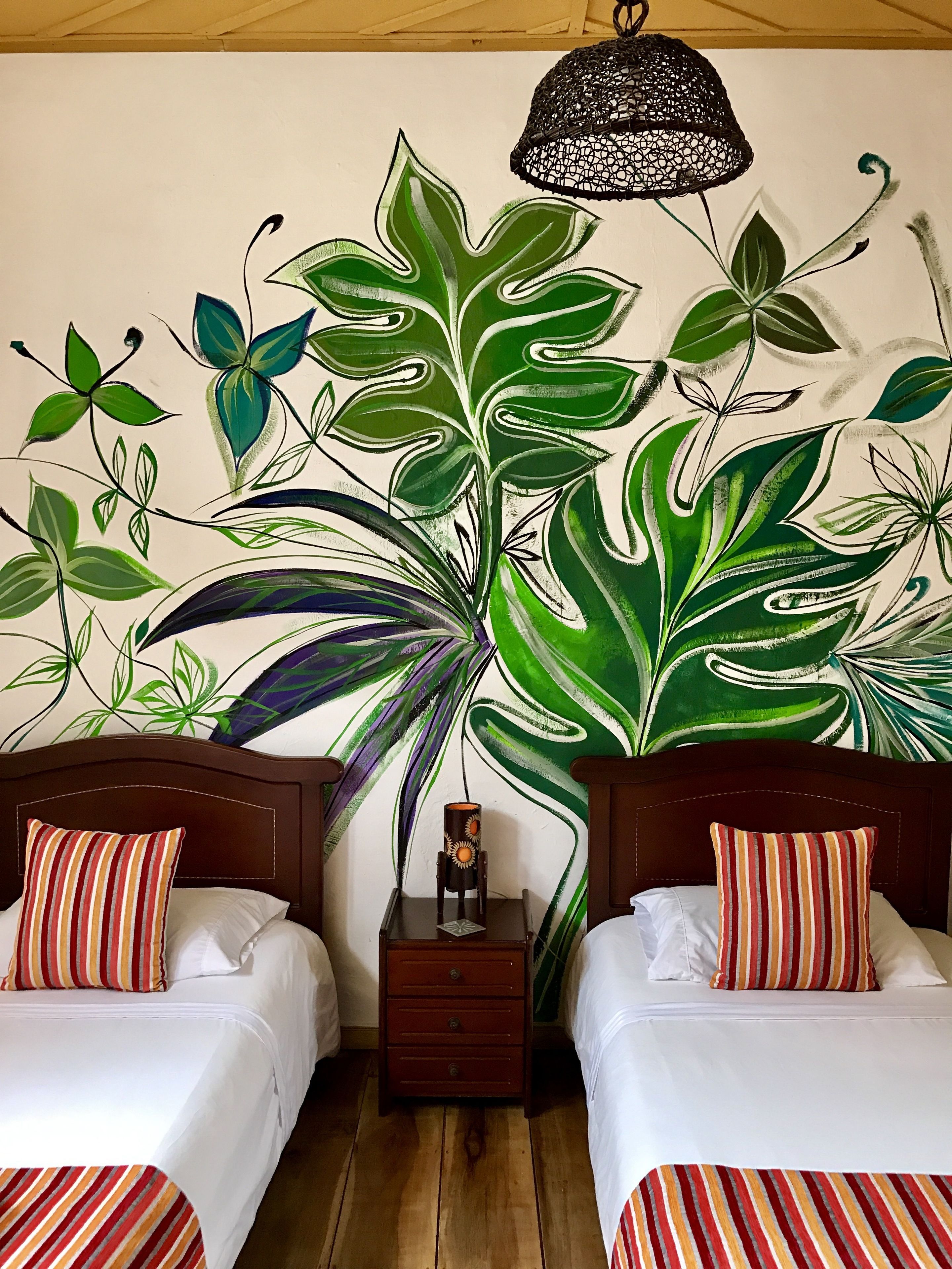 A room with two beds, a nightstand, and a large wall mural of tropical leaves.