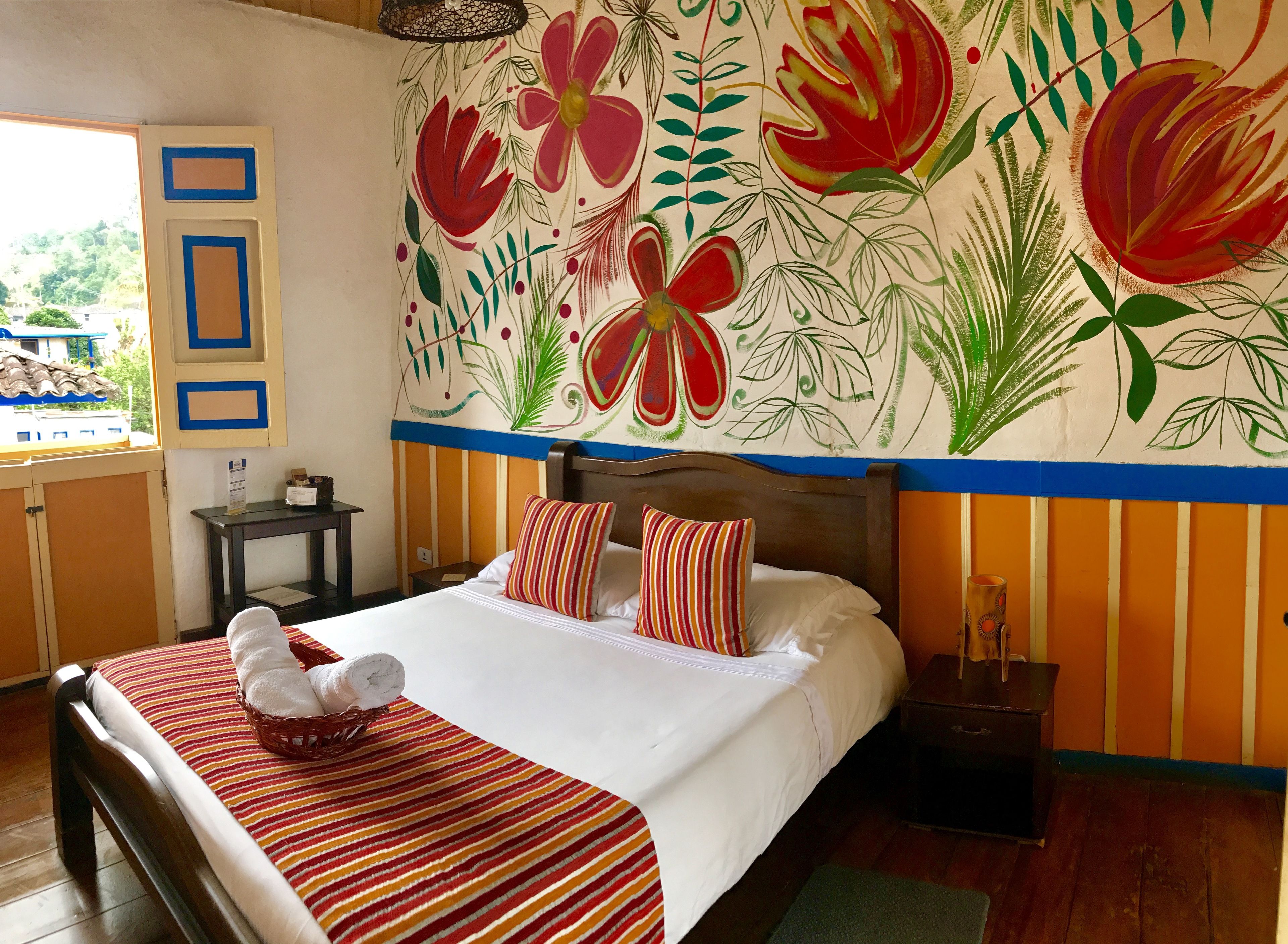 A hotel room with a bed, pillows, a nightstand, a wall mural, and a window.