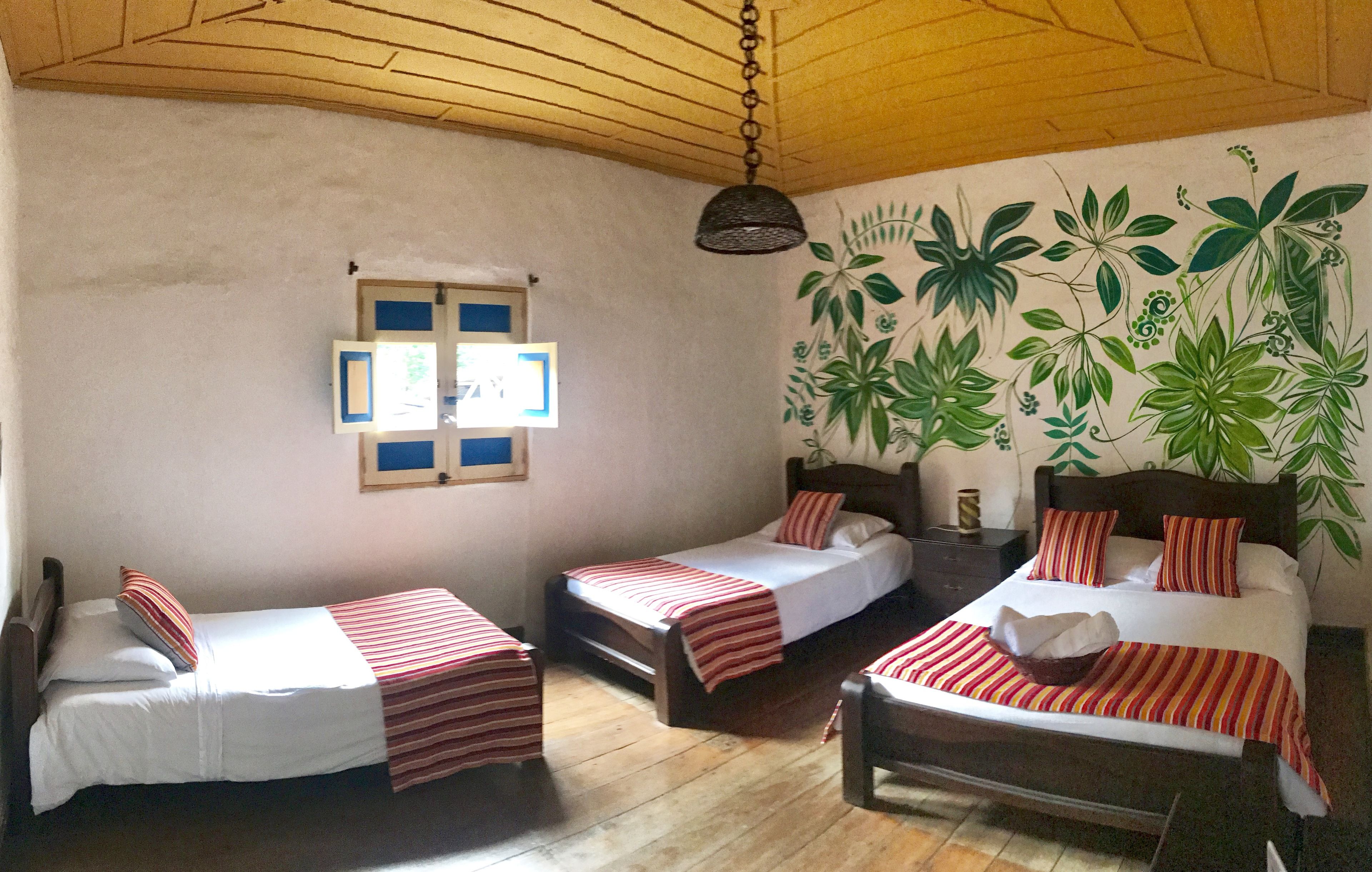 Habitación con tres camas individuales, suelo de madera, lámpara colgante y un mural en la pared con follaje verde.