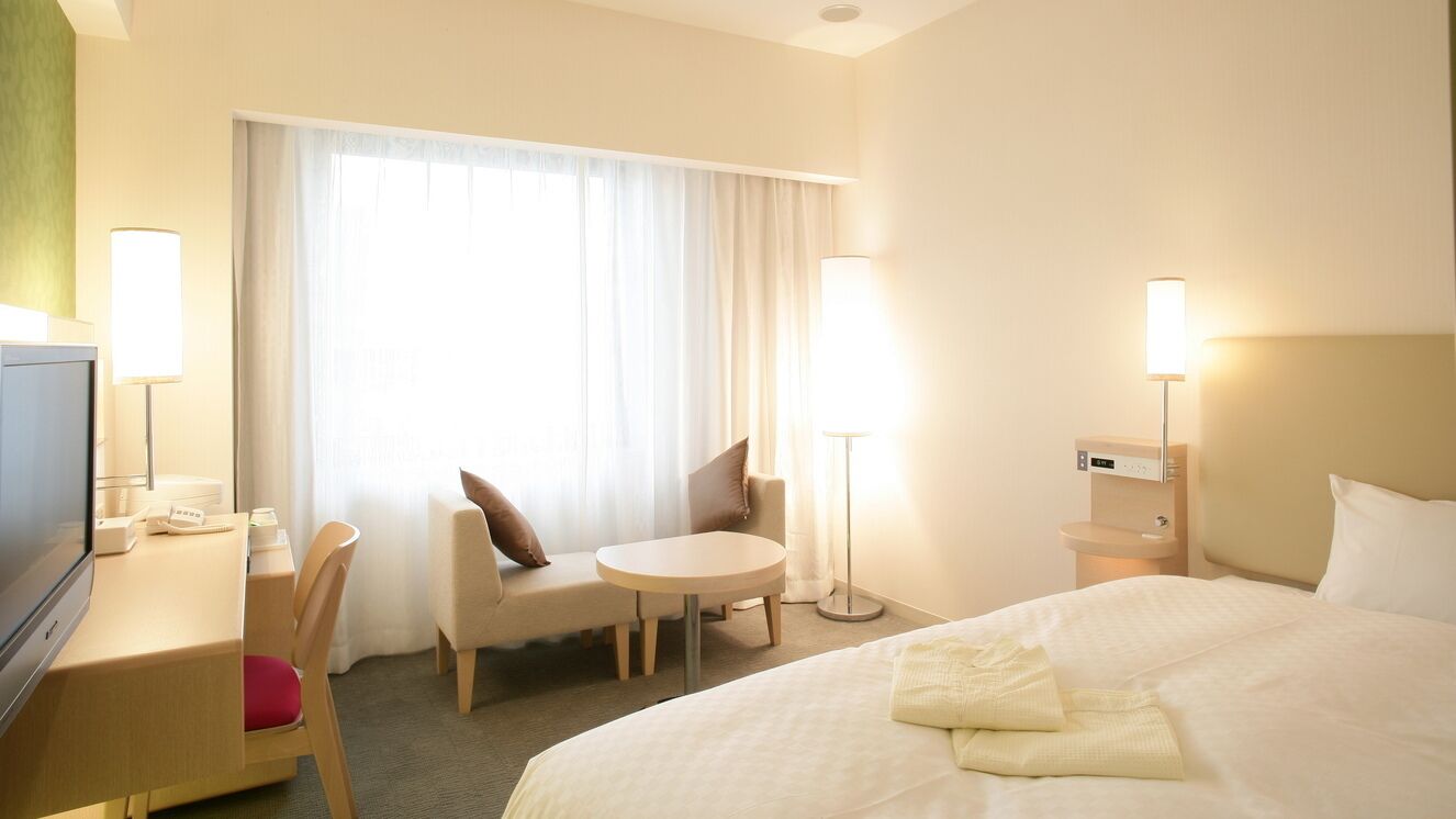 Une chambre d’hôtel comprenant un lit, un bureau, une chaise, une table ronde, un téléviseur et une fenêtre avec des rideaux.