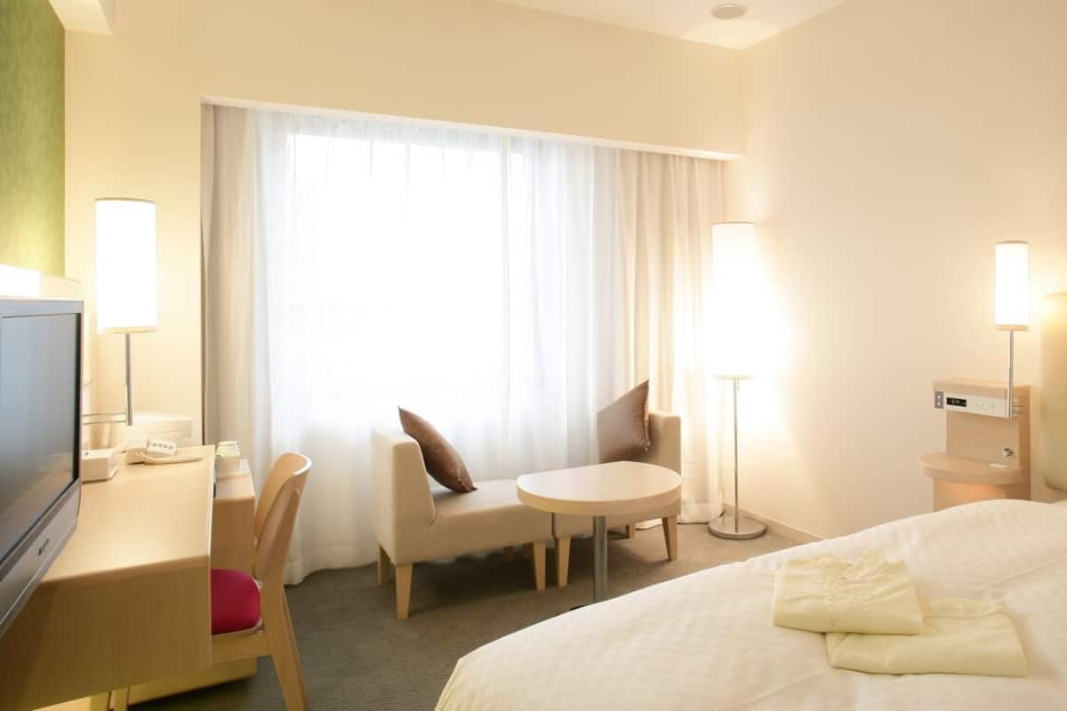 A hotel room with a bed, a desk, a chair, a round table, a TV, and a window with curtains.