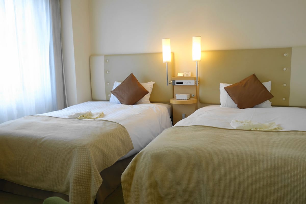 Two adjacent hotel beds with headboards, each with a pillow and a folded towel. There is a nightstand with a lamp between the beds.