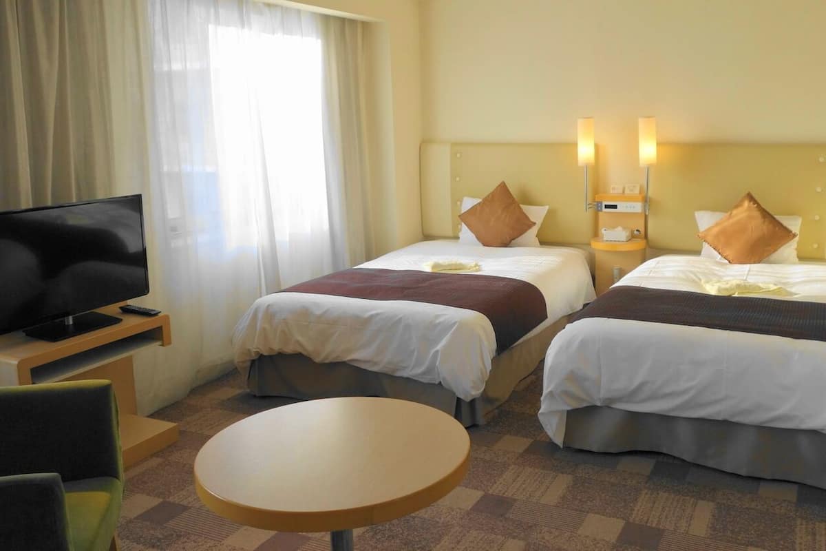 A hotel room with two beds, a TV, a small table, and a window with curtains.