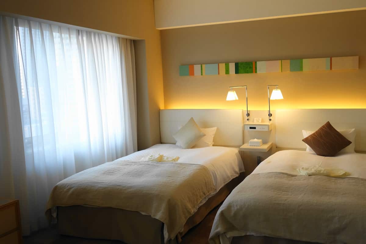 A hotel room with two beds, a nightstand with a lamp, and a wall-mounted control panel.