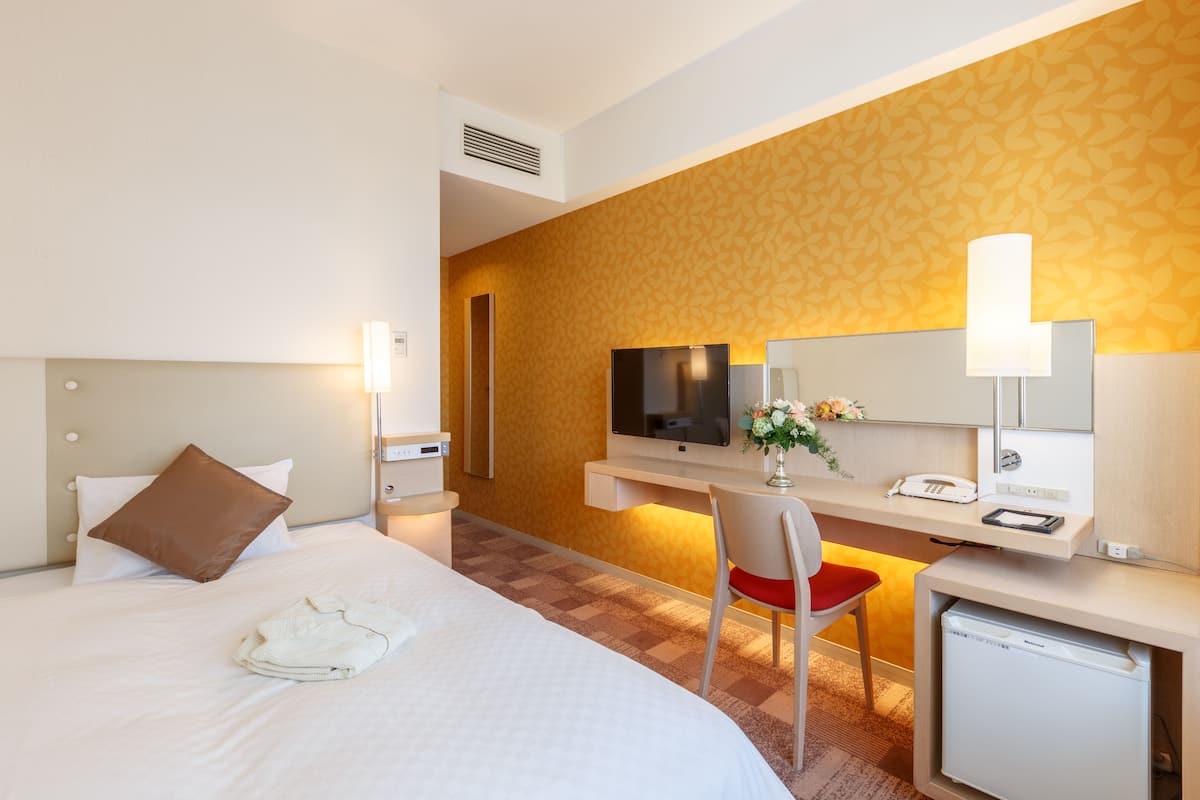A hotel room with a bed, a desk with a chair, a television, a mirror, and a small refrigerator.
