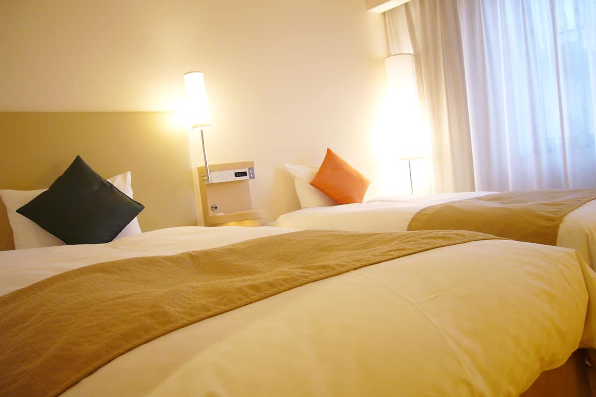 A hotel room with two beds, a nightstand with a lamp, and a window with curtains.