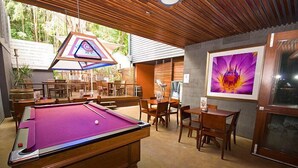 A pool table in a room with wooden furniture, a framed painting, and a view of greenery outside. - Global Backpackers - Port Douglas (Port Douglas)