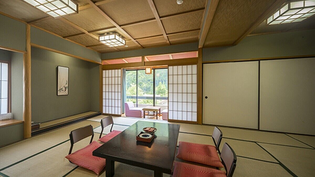 Um quarto japonês tradicional com piso de tatame, uma mesa baixa com almofadas, portas deslizantes e vista para o verde lá fora.
