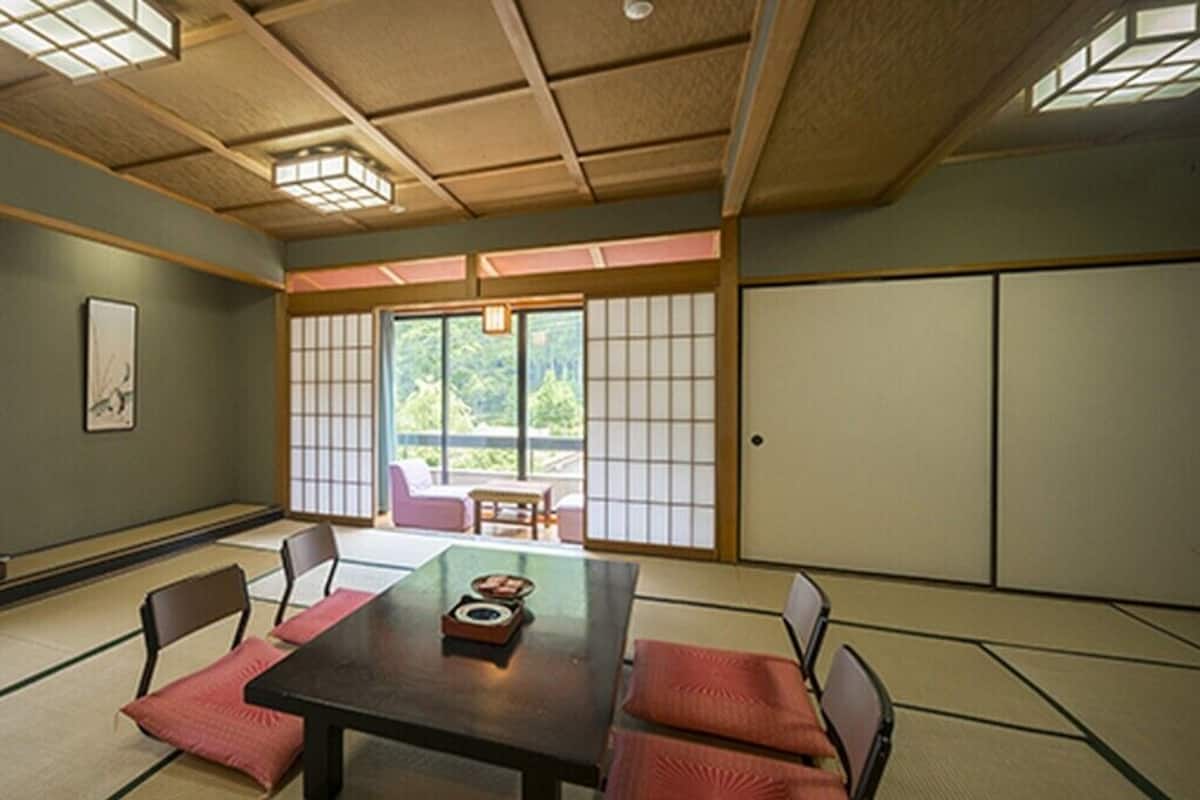 A traditional Japanese room with tatami flooring, a low table with cushions, sliding doors, and a view of greenery outside.