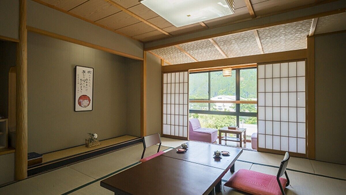 Um quarto japonês tradicional, com piso de tatame, uma mesa baixa com almofada, uma porta deslizante com vista e uma pequena mesa com uma cadeira.