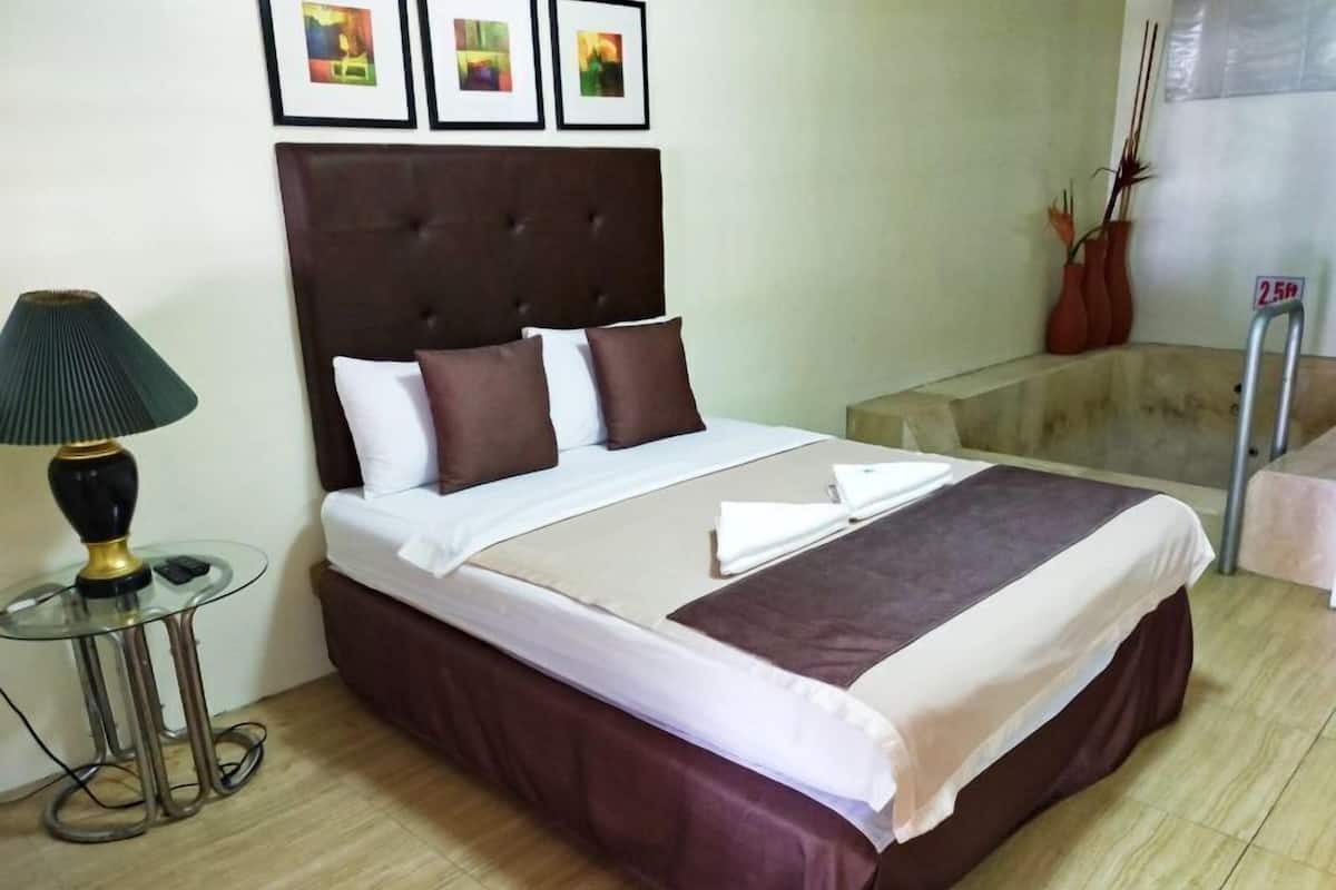 A neatly made bed with a brown headboard, white and brown pillows, and a beige bedspread. There is a glass table with a lamp beside the bed.