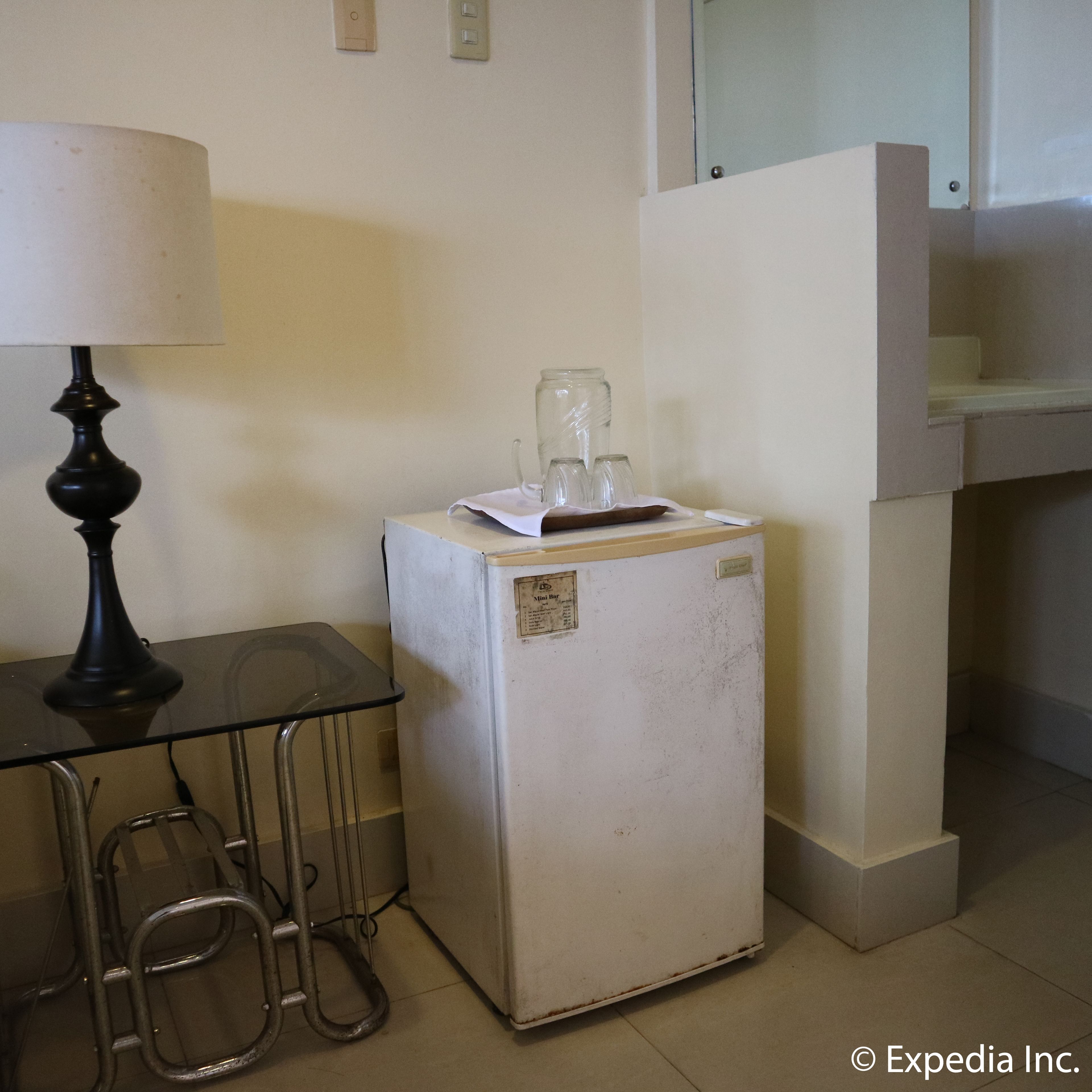 A small kitchenette with a refrigerator, a table with a lamp, and a chair.