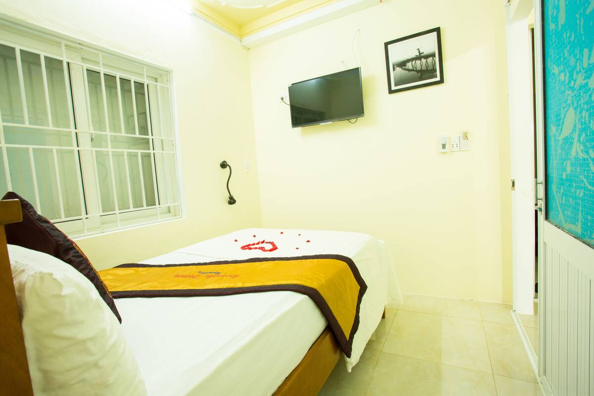 A single bed with a white and yellow bedspread, a television mounted on the wall, and a window with blinds.